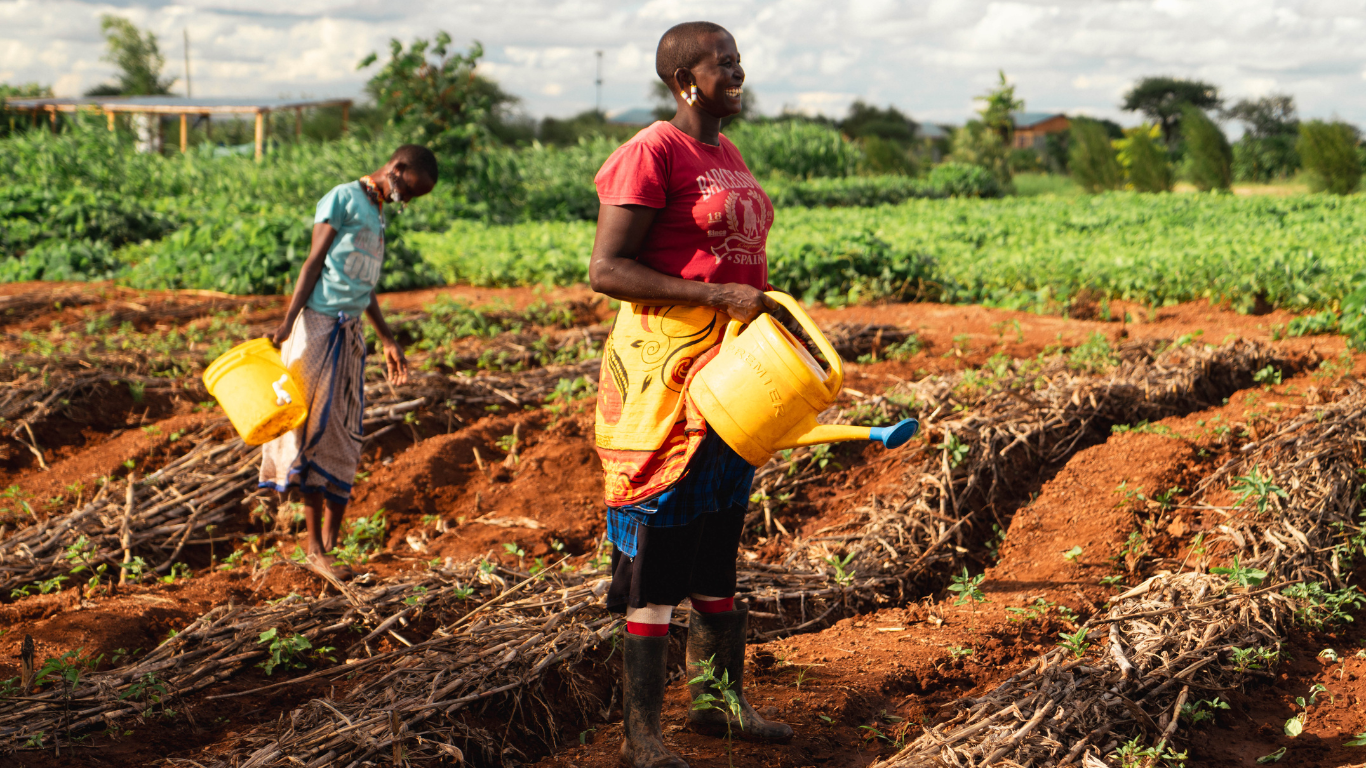 A picture of 2 women workers on Ambokili Farm smiling holding watering cans. It illustrates their happiness when working and demonstrates how Ambokili Farm is truly investing in Employee well-being